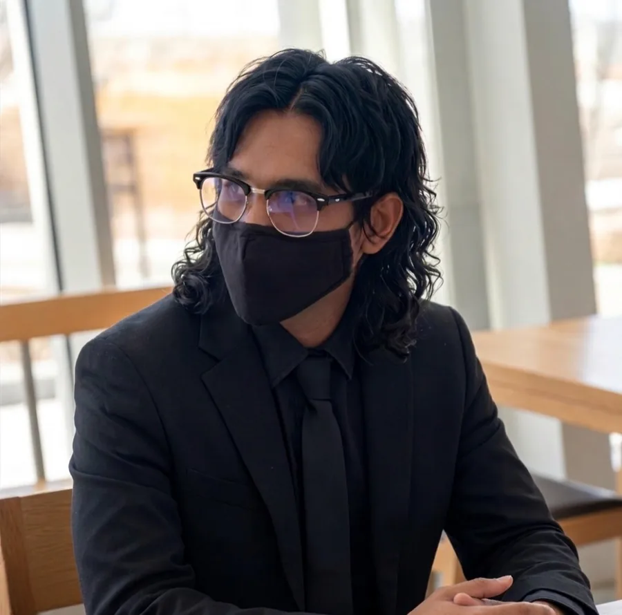 Portrait: a person with dark wavy hair, glasses and a black face mask, wearing a black suit and tie